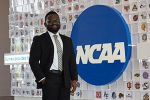 An African American man stands in front of an NCAA logo. Tiles featuring logos of various teams decorate the wall behind him.
