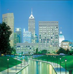 A rendering of Indianapolis at dusk. Thin, black lampposts line a canal that runs toward gray skyscrapers.