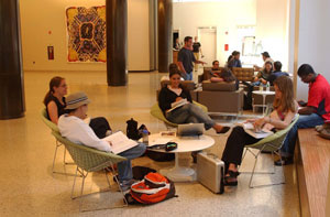 Students are seated in clusters at an open study area in a large hallway.