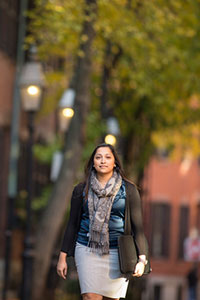 Female student walking, with a briefcase bag slung over her shoulder. In the background are lit lampposts, deciduous trees, and brick buildings with black shutters.