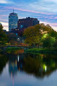 Boston at dusk. Pink and blue stratus clouds spread across the sky. In the foreground, trees and a skyscraper are reflected on a river. A footbridge stretches over a narrow portion of the river.