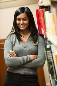A female student leans against a shelf of library books, smiling.