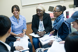Inside a classroom, diverse students sit in a circle, listening and smiling, as one student speaks. Each student holds a pen and a pile of papers.