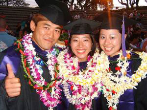 Three graduates wearing leis—1 male, 2 female—pose after the ceremony, smiling. One graduate is giving a 'thumbs up.'