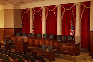 A moot courtoom. Five red curtains, trimmed in gold, hang behind the bench. Two American flags also stand behind the bench.