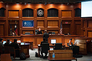A student stands before the bench during a mock trial. A blue sign, embedded into the wall behind the bench, says, 'St Mary's University School of Law.' An American flag and two other flags also stand behind the bench