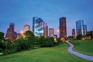 Houston at night. A winding footpath cuts through a grassy field, toward a backdrop of skyscrapers. A patch of deciduous trees stand to the left of the path.