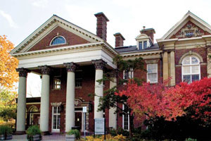 Flavelle House—a decorative brick building with white accents. Four white columns lead to the main entrance. Deciduous trees and shrubs are in view.