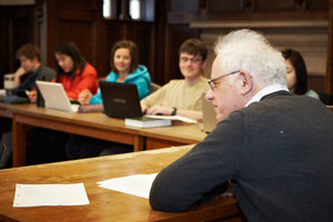 A professor and students are seated at an angled wooden table. Sheets of paper are scattered in front of the professor, who is looking off to his left. Laptops are open in front of the students. Several students look at their laptops, several toward the professor.