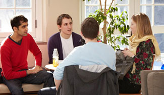 In a well-lit study area, four students sit in a circle of chairs and speak. A potted tree stands behind the students.