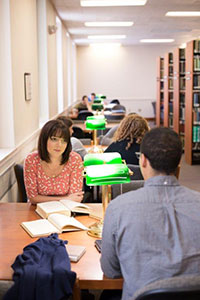 In the library, a female student listens as the male student sitting across from her speaks. Two books are opened in front of her. Other students occupy the row of study tables behind her. Two green desk lamps sit on each table.