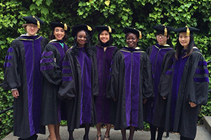 Diverse graduates, wearing black robes with purple accents, pose on a stairway platform outdoors.