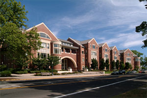 Angled view of law school exterior. A brick building with beige accents that sits a few feet away from a two-lane road. An arch leads into the school's main entrance. Deciduous trees are in view.