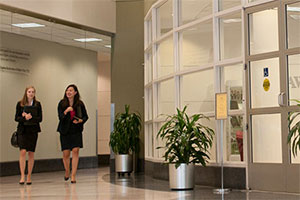 Two women walk down a curved hallway. Potted plants are to their left, outside of a room with glass walls.