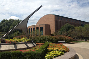 A modern sculpture, surrounded by flowers, points toward the cloudy sky. In the background, the law school building is made of brick and has a mixture of curved and angled edges. Many deciduous trees are in view.