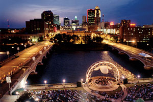 Des Moines at night. The city is brightly lit. An event is happening on an outdoor stage along the water. Many people are in attendance.