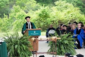 A man speaks from a podium during commencement. Faculty members are seated behind him, listening to the speech.